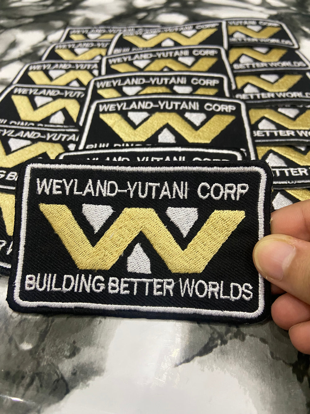 Patches with 'Weyland-Yutani Corp' logo and text held by a hand.