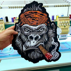 Embroidered patch of a gorilla smoking a cigar held in front of an embroidery machine.
