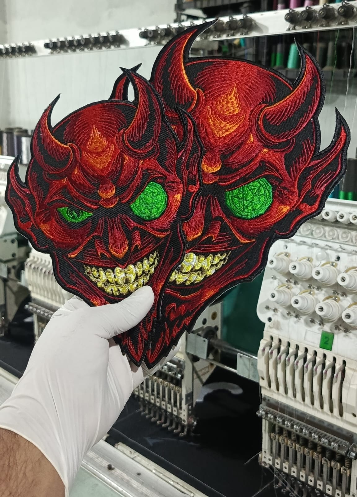 cyberpunk devil illuminati patches. red illuminati symbols

A hand wearing a white glove holds up a large embroidered patch featuring two red devil faces with glowing green eyes and yellow teeth, with an industrial embroidery machine in the background.